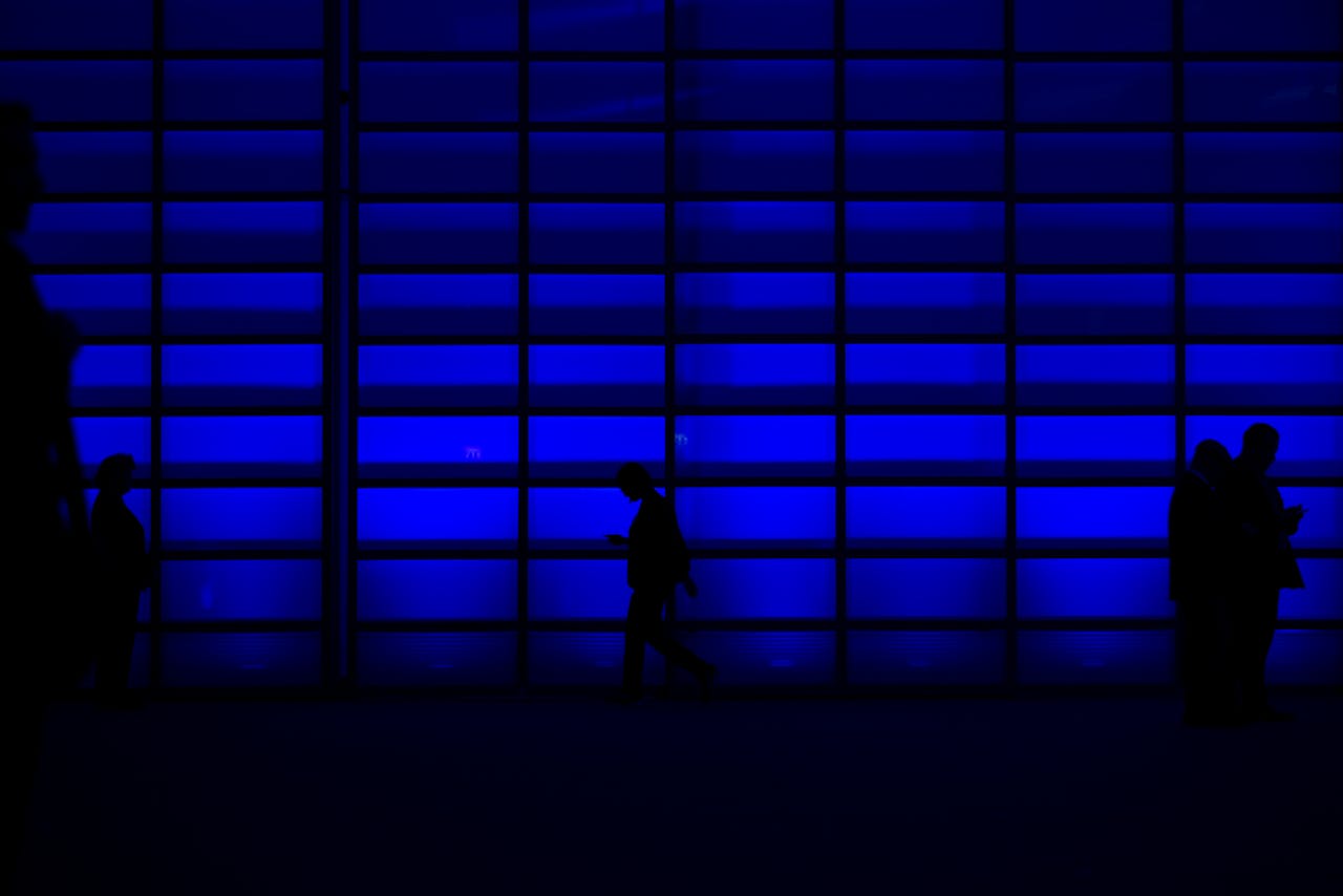 Silhouettes of people walking and using phones against a blue illuminated wall.